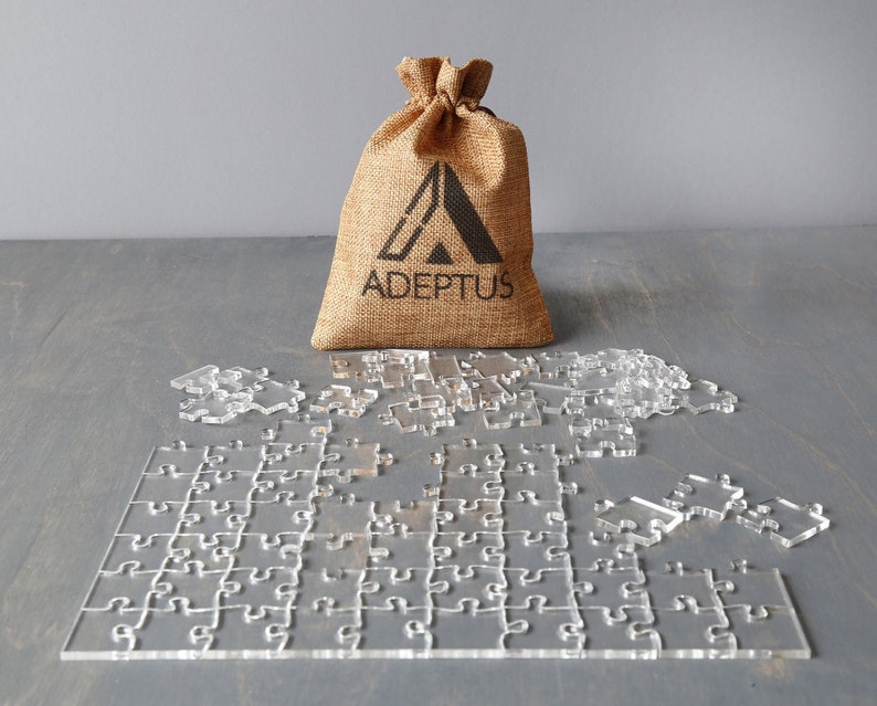 May include: A clear acrylic jigsaw puzzle with a burlap bag that says "ADEPTOS" on it. The puzzle pieces are scattered around the bag.