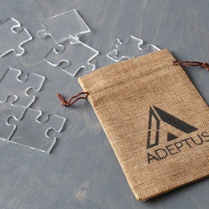 May include: A burlap pouch with a drawstring closure and a black logo that says "ADEPPTUS" with a stylized triangle design. The pouch is sitting on a gray surface with clear plastic puzzle pieces scattered around it.