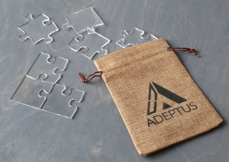May include: A burlap pouch with a drawstring closure and a black logo that says "ADEPPTUS" with a stylized triangle design. The pouch is sitting on a gray surface with clear plastic puzzle pieces scattered around it.