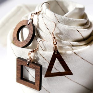 May include: Three wooden earrings with copper-colored hooks. One earring is a circle, another a square, and the third a triangle. The earrings are displayed on a white fabric with thin brown stripes.