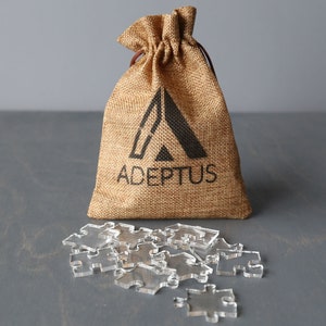 May include: A burlap bag with a drawstring closure and the word "ADEPTUS" printed on it. The bag is filled with clear acrylic puzzle pieces.