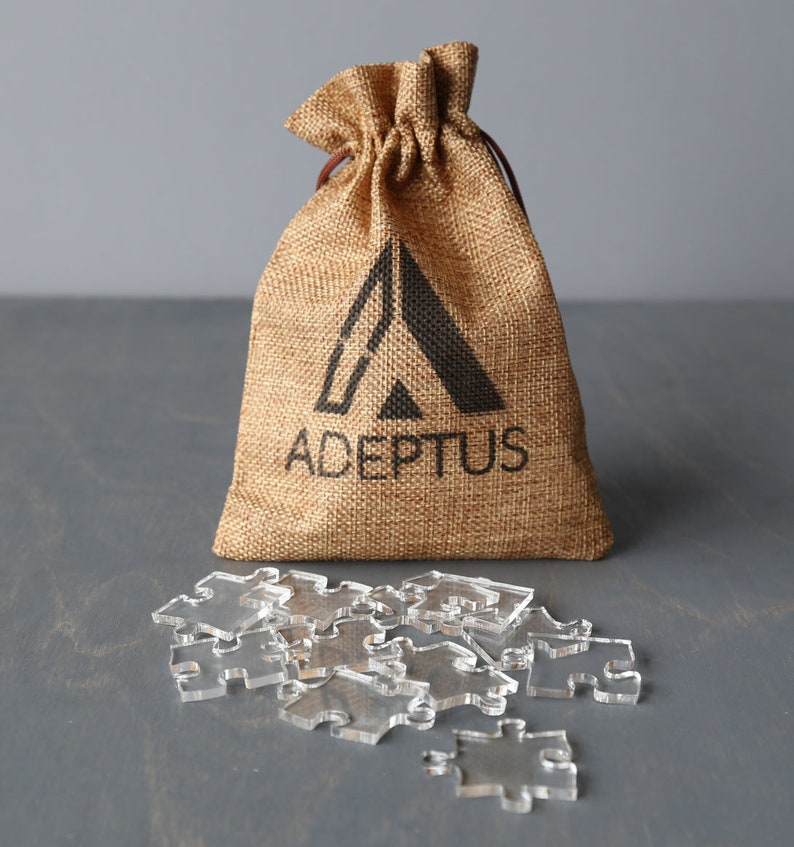 May include: A burlap bag with a drawstring closure and the word "ADEPTUS" printed on it. The bag is filled with clear acrylic puzzle pieces.