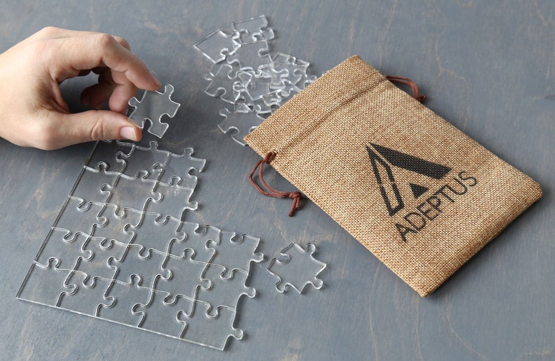May include: A clear acrylic jigsaw puzzle with a burlap bag that has the logo "ADEPTOS" printed on it.