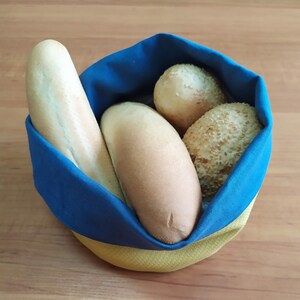 May include: A blue and yellow fabric bread basket filled with various types of bread. The basket contains a baguette, two rolls, and a sesame seed bun. The basket is made of blue and yellow fabric.