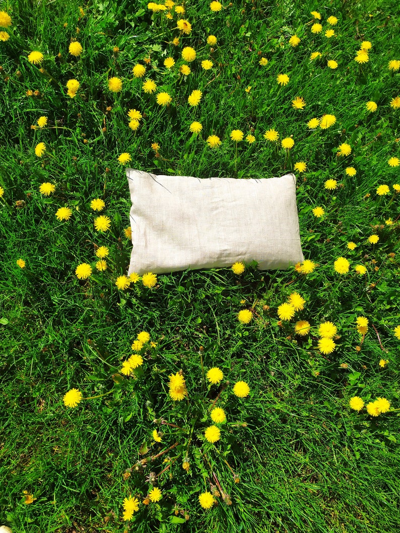 May include: A white linen pillow resting on a bed of green grass with yellow dandelions.