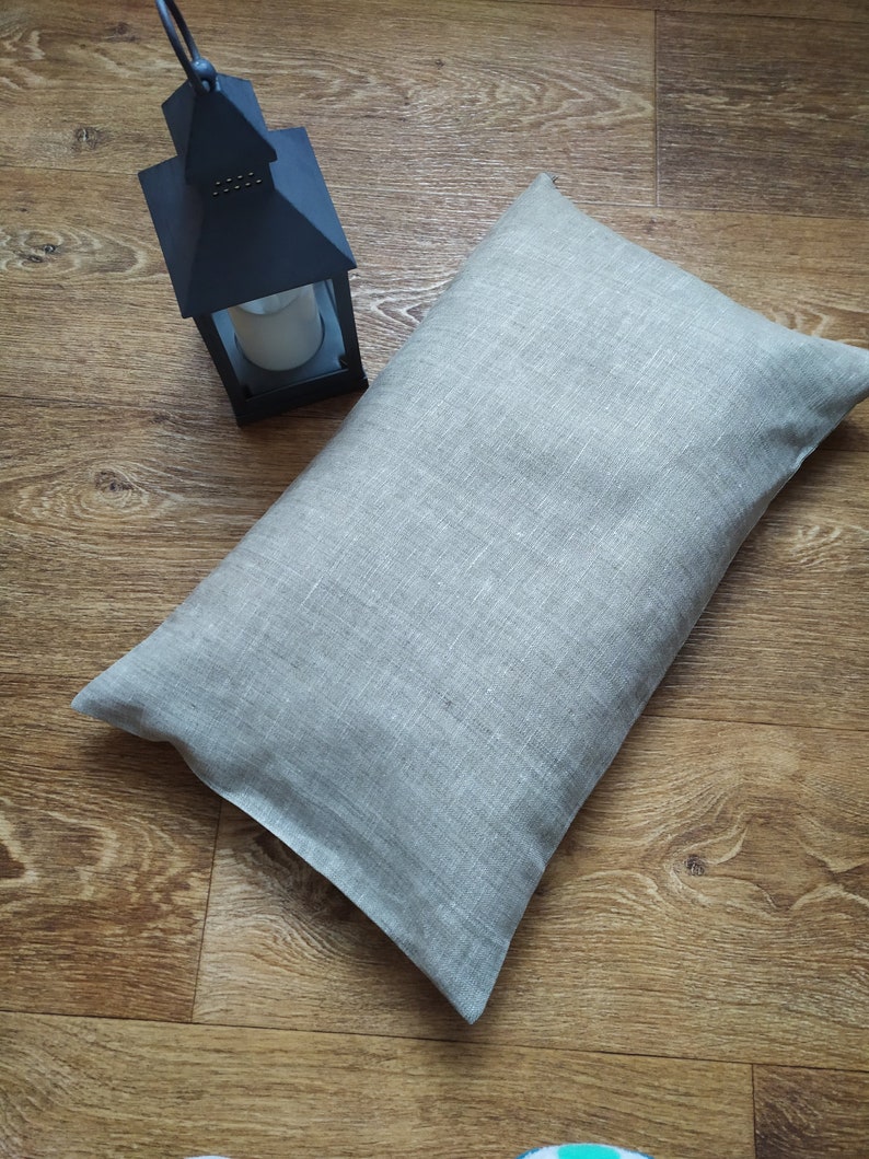 May include: A light gray linen pillow with a subtle texture. The pillow is lying on a wooden floor.
