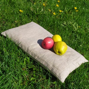 May include: A light gray linen pillow with three apples on top. Two of the apples are green and one is red. The pillow is lying on a bed of green grass.