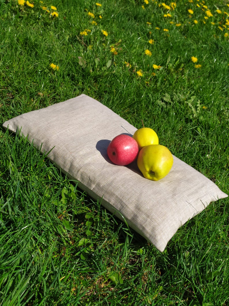 May include: A light gray linen pillow with three apples on top. Two of the apples are green and one is red. The pillow is lying on a bed of green grass.