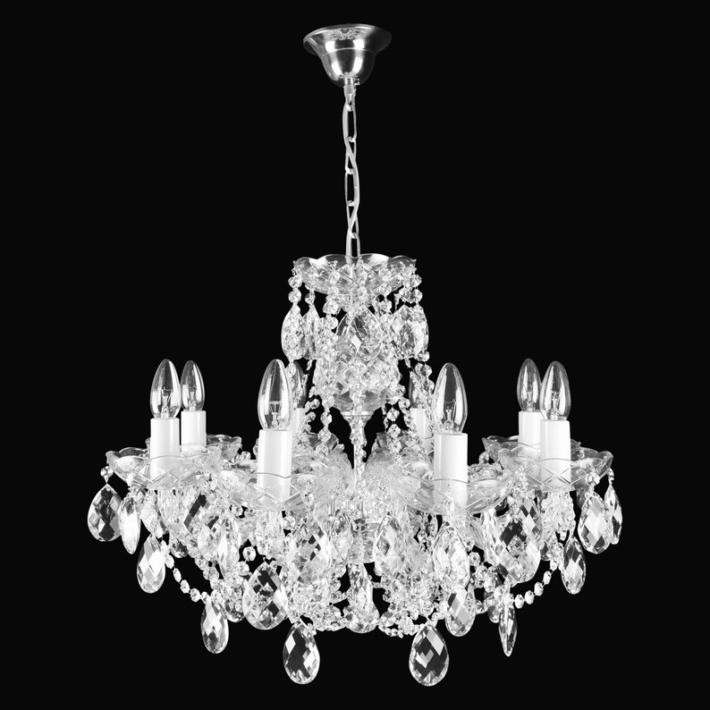 May include: A crystal chandelier with a silver finish and clear glass crystals. The chandelier has six light bulbs and a chain hanging from the ceiling.