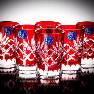 Könnte beinhalten: Satz mit sechs roten Kristall-Schnapsgläsern mit geschliffenem Glasdesign. Jedes Glas hat einen weißen Rand und ein blaues Etikett mit dem Text "Crystal Bohemia" und einem Logo.