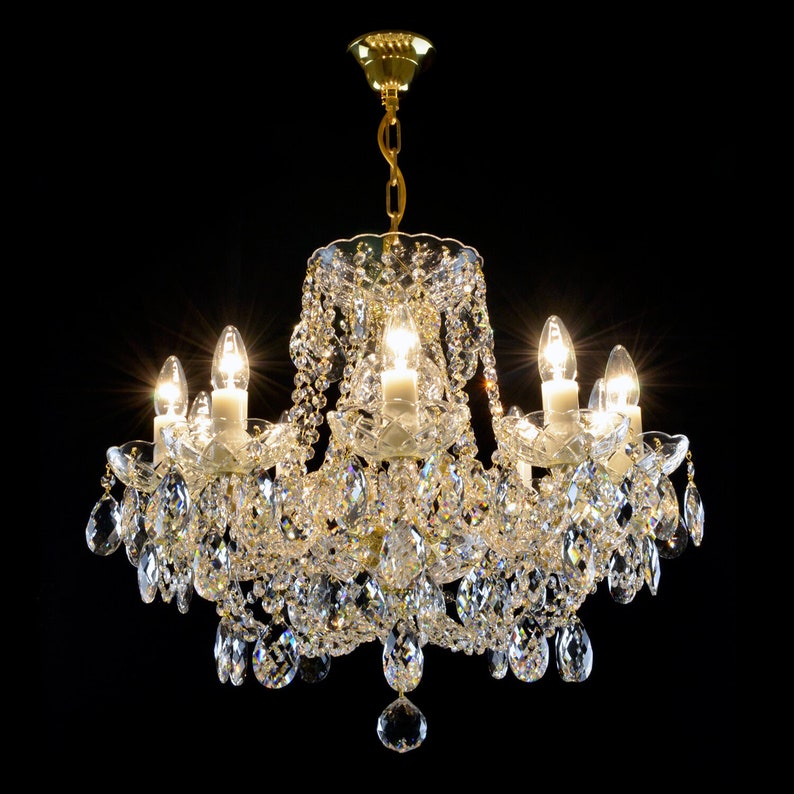 May include: A crystal chandelier with a gold finish and multiple arms, each with a clear glass candle holder and a light bulb. The chandelier is adorned with numerous crystal drops, creating a sparkling effect.