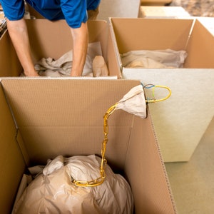 May include: A person packing a brown paper bag into a cardboard box. The bag is connected to a gold chain that is hanging out of the box. The box is labeled "SP-91".
