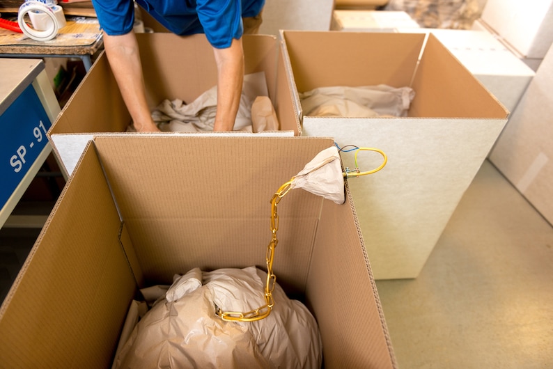 May include: A person packing a brown paper bag into a cardboard box. The bag is connected to a gold chain that is hanging out of the box. The box is labeled "SP-91".