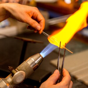 May include: A glassblower uses a torch to heat a glass rod held with tweezers. The torch has a blue flame and the glass rod is glowing orange.