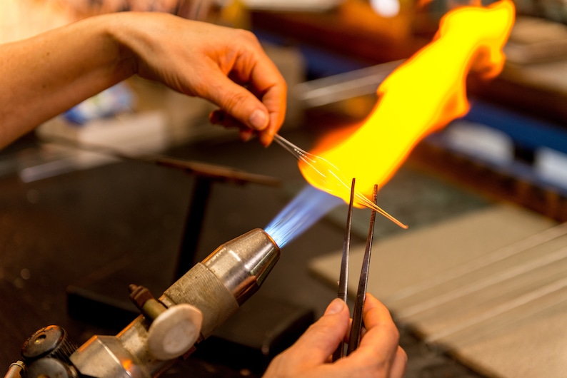 May include: A glassblower uses a torch to heat a glass rod held with tweezers. The torch has a blue flame and the glass rod is glowing orange.