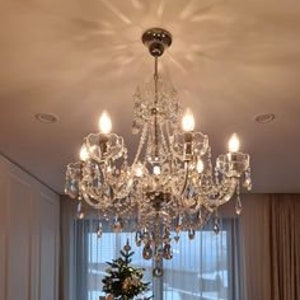 May include: A crystal chandelier with a silver finish and clear crystal drops. The chandelier has six lights and is hanging from a white ceiling.