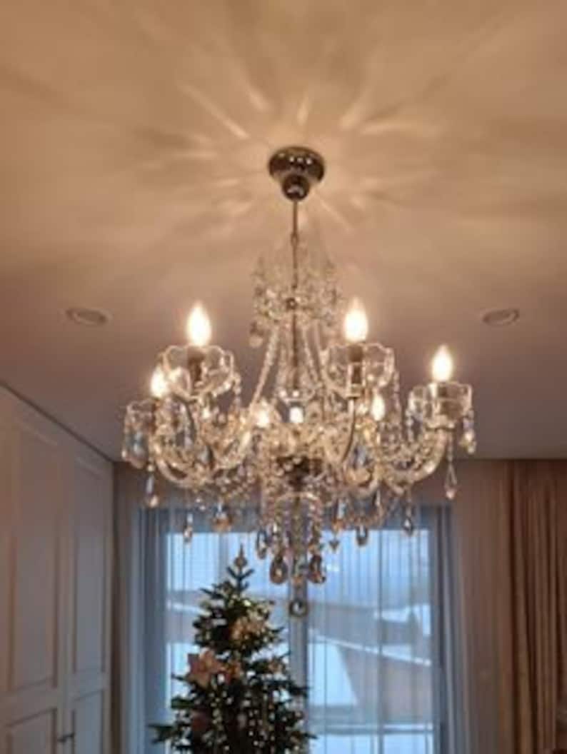May include: A crystal chandelier with a silver finish and clear crystal drops. The chandelier has six lights and is hanging from a white ceiling.