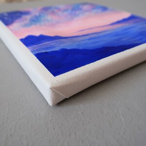 May include: A small canvas painting with a pink and blue sky and blue mountains in the foreground.