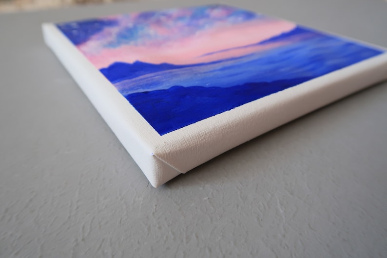 May include: A small canvas painting with a pink and blue sky and blue mountains in the foreground.