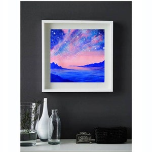 May include: A framed painting of a pink and blue landscape with a starry sky and a body of water. The painting is in a white frame and is hanging on a dark gray wall.