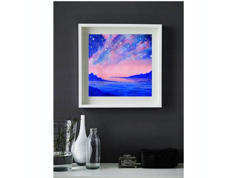 May include: A framed painting of a pink and blue landscape with a starry sky and a body of water. The painting is in a white frame and is hanging on a dark gray wall.