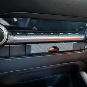 May include: A gray, textured, rectangular cover piece installed over the hazard light button on a vehicle's climate control console. The console features black buttons, a digital display, and two circular knobs labeled AUTO and SYNC.