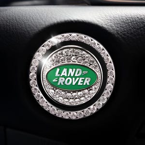 May include: A silver and crystal-studded car ignition button cover. The cover features the Land Rover logo in green and white. The button is surrounded by a ring of clear crystals, adding a touch of luxury to the vehicle's interior.