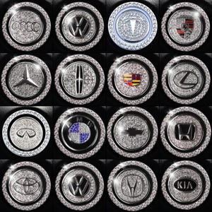 May include: A collection of sixteen car emblem accessories, each featuring a different brand logo. The emblems are circular, with a silver-colored, gem-studded border. The logos are also silver and gem-studded, set against a black background.