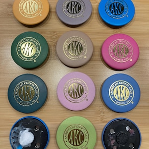 May include: A collection of twelve colorful round objects, each featuring the text "AMERICAN KENNEL CLUB FOUNDED 1884" in gold lettering. The objects are in various colors, including orange, blue, green, and pink.