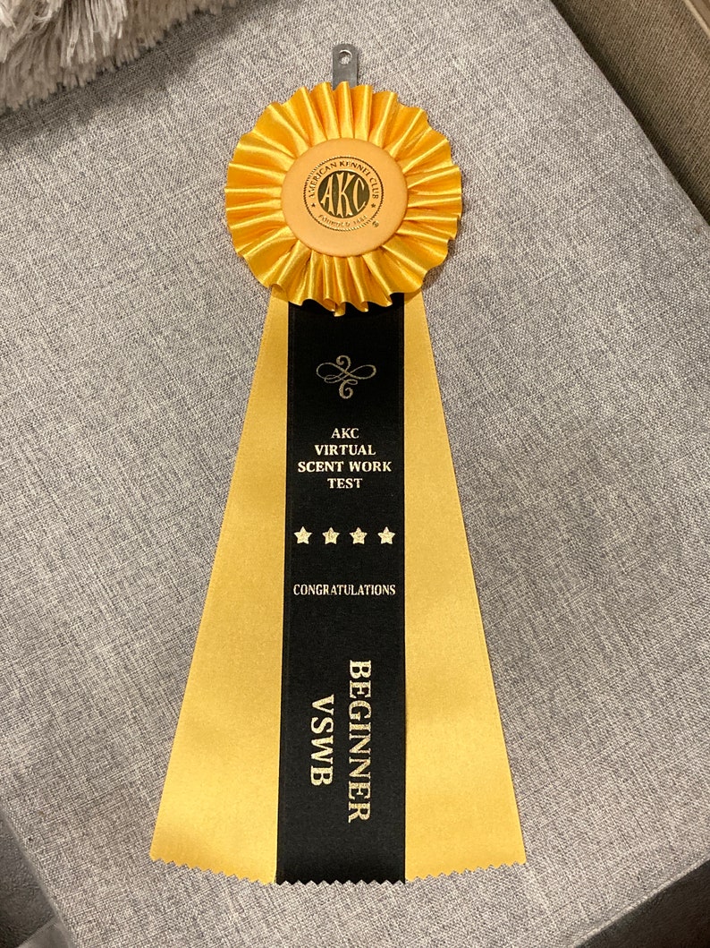 May include: A gold ribbon with a black stripe down the centre. The ribbon has the text "AKC Virtual Scent Work Test" and "Congratulations Beginner VSWB". The ribbon is tied with a gold rosette with the text "American Kennel Club" in the centre.