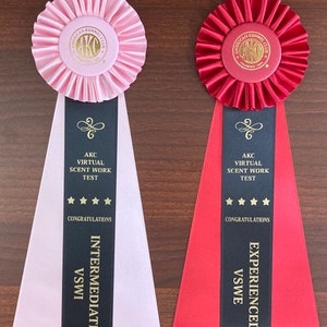 May include: Two pink and red ribbons with black stripes and gold lettering. The ribbons are for the American Kennel Club Virtual Scent Work Test. The pink ribbon is for the Intermediate level and the red ribbon is for the Experienced level.