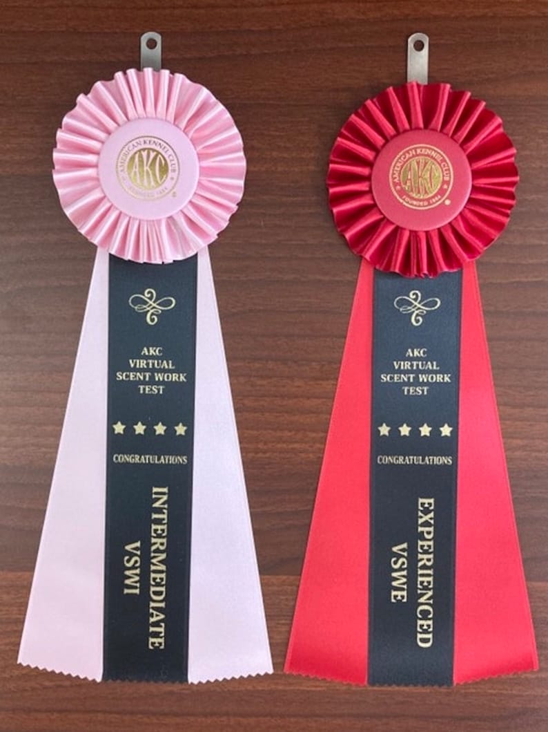 May include: Two pink and red ribbons with black stripes and gold lettering. The ribbons are for the American Kennel Club Virtual Scent Work Test. The pink ribbon is for the Intermediate level and the red ribbon is for the Experienced level.