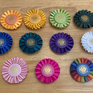May include: A collection of 15 colourful rosettes with gold lettering. The rosettes are made of satin ribbon and have a ruffled edge. The lettering on the rosettes reads "AKO".
