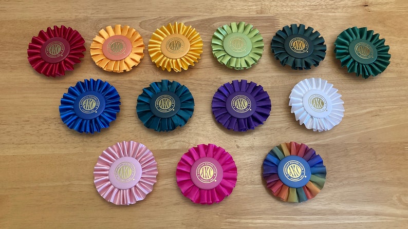 May include: A collection of 15 colourful rosettes with gold lettering. The rosettes are made of satin ribbon and have a ruffled edge. The lettering on the rosettes reads "AKO".