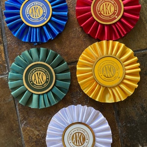 May include: Five rosette ribbons in blue, red, green, yellow, and white. Each ribbon features a gold circle with the text "American Kennel Club" and "AKC" in the centre. The text "Founded 1884" is below the "AKC" text.