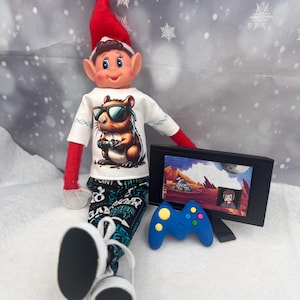 May include: An elf doll wearing a white t-shirt with a cartoon groundhog wearing sunglasses and holding a game controller. The elf is also wearing black and teal patterned pants, a red elf hat, and white sneakers. A small TV and blue game controller are nearby.