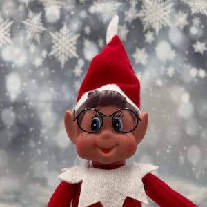 Könnte beinhalten: Eine rote und weiße Elfenpuppe mit braunen Haaren, die eine schwarze Brille und eine rote Mütze trägt. Der Elf lächelt und hat blaue Augen. Der Hintergrund ist ein verschwommenes Bild von weißen Schneeflocken.