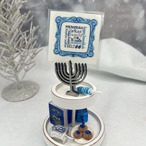 May include: A two-tiered white wooden tray featuring a Hanukkah sign with blue text, a gray metal menorah, a blue and white striped lollipop, a blue book, a silver dreidel, and a small plate with three brown cookies with blue centers.