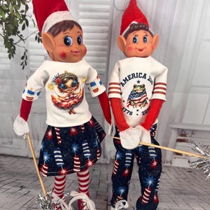 May include: Two elf dolls wearing patriotic outfits. One doll wears a white shirt with a graphic and a blue skirt with firework print. The other doll wears a white shirt with "America 202" and a frog graphic, and blue pants with firework print. Both wear red elf hats.