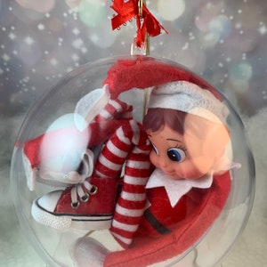 May include: A red and white striped elf doll wearing red sneakers is sitting inside a clear glass ball ornament. The ornament has a gold top and a red bow.