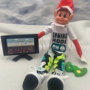 May include: A Christmas elf doll wearing a white shirt with the text "Gaming Mode On" in green and blue letters, green and black patterned pants, and white sneakers. The elf is holding a green video game controller and sitting in front of a small black television with a video game on the screen.