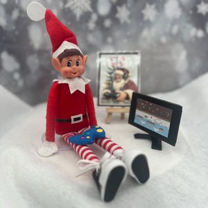 May include: A Christmas elf doll wearing a red outfit with a white collar and hat, and striped legs. The elf is holding a blue video game controller and sitting near a small TV and a painting of Santa Claus.