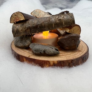 May include: A small, lit candle sits on a wooden log slice surrounded by small pieces of driftwood. The scene is set on a white, fluffy surface.