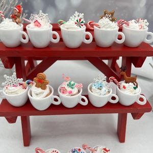 Könnte beinhalten: Eine Sammlung von Miniatur-Tassen aus weißer Keramik, gefüllt mit künstlicher Schlagsahne und verziert mit verschiedenen weihnachtlichen Anhängern wie Lebkuchenmännchen, Rentieren, Zuckerstangen und Schneeflocken auf einem roten Holzständer.