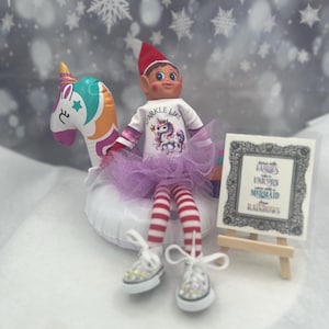 May include: An elf doll wearing a red and white hat, a white shirt with a unicorn graphic, and a purple tutu. The elf is sitting on a unicorn-shaped inflatable float. The elf has red and white striped legs and sparkly white sneakers. A small sign on an easel reads "Fairies Unicorn Mermaid Rainbows."