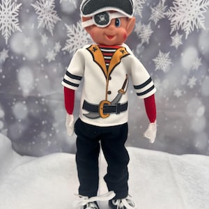 May include: A doll dressed as a pirate, wearing a black and white pirate hat with a skull and crossbones, an eye patch, and a white jacket with gold trim. The doll is wearing black trousers, red sleeves, and white trainers.