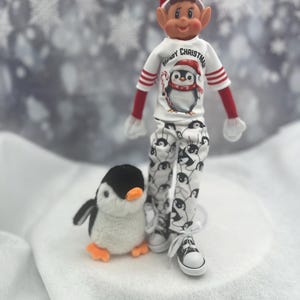 May include: An elf doll in a red hat, a white shirt with a penguin graphic and the text "Merry Christmas", and penguin-patterned trousers. A plush penguin toy sits beside the elf. The elf is wearing black and white trainers.