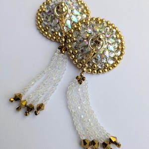 May include: Two decorative nipple pasties. Each pastie is round, with a gold beaded border and a central arrangement of clear and iridescent crystals. White beaded tassels with gold accents hang from each pastie.