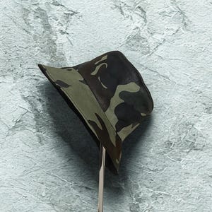 May include: A camouflage bucket hat in shades of green, brown, and black. The hat is displayed on a silver stand against a textured, light blue background. The hat's brim is a solid green color.