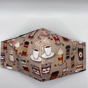 May include: A beige fabric face mask with a coffee theme. The mask is covered in a repeating pattern of coffee cups, coffee beans, and coffee-related words like "Espresso" and "Latte".